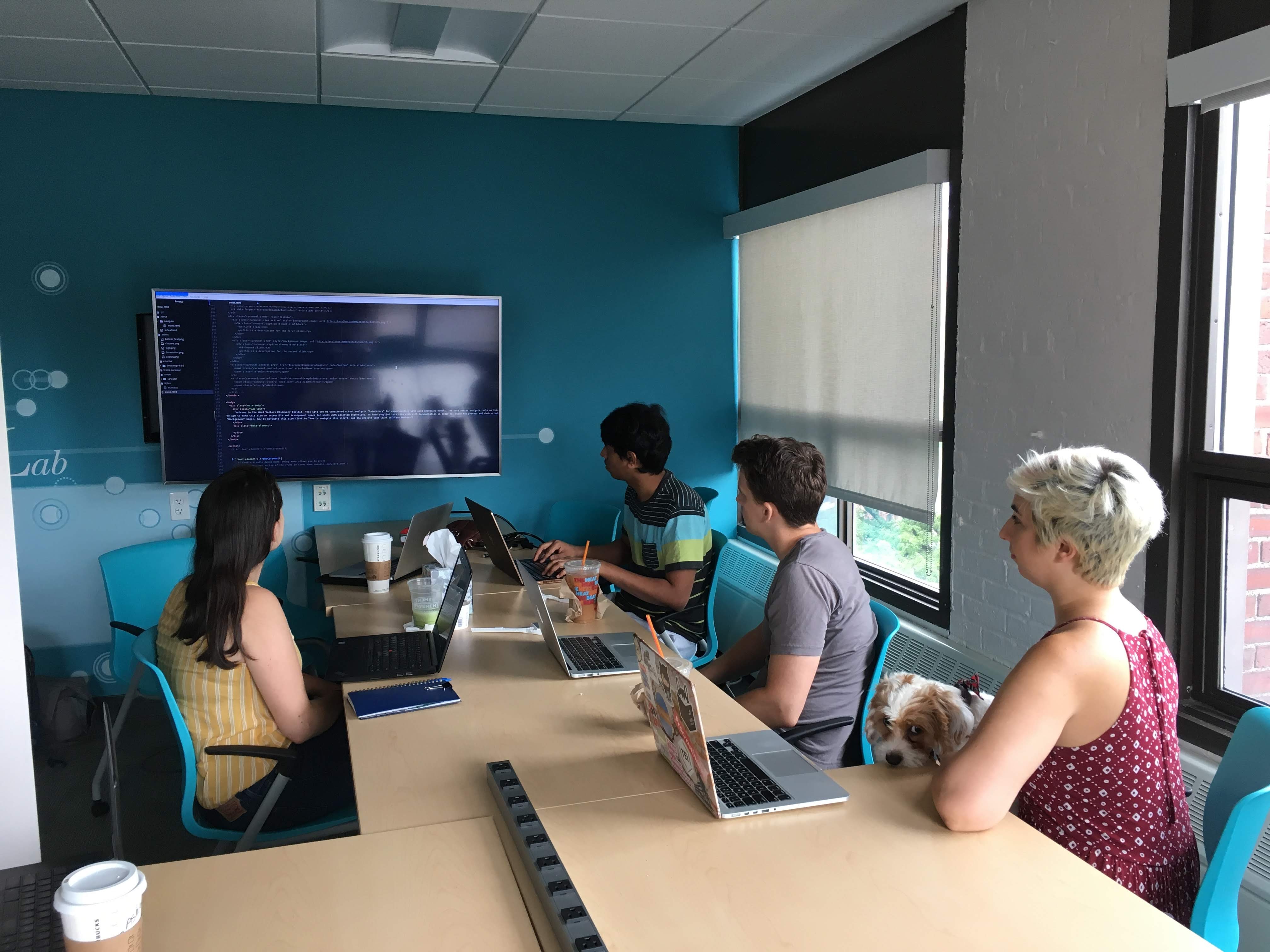 Four people face a monitor on a wall, examining some code.